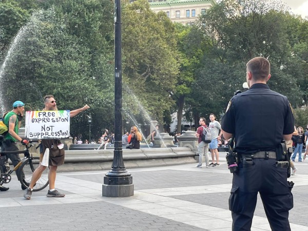 A man with a beard and sunglasses is wearing an orange brown and white shirt, and is yelling at the camera. He holds a sign labeled “Free Expression Not Suppression” in front of the fountain at Washington Square Park. There are people standing and sitting beside the fountain in the background.