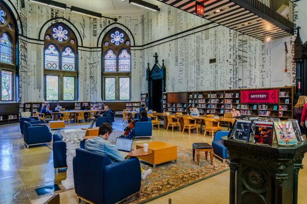 The interior of Mulberry Street Library with a shelf of magazines and books and several pots of plants on top of it. There is a person reading at a table behind the bookshelf. There are more bookshelves by the walls.