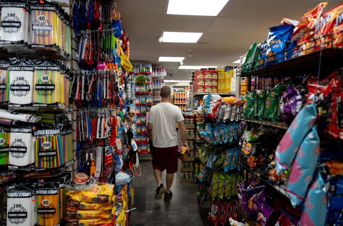 A man dressed in a white shirt with red shorts and black shoes walks down the middle of rows of grocery items, including chips, straws, and toys.