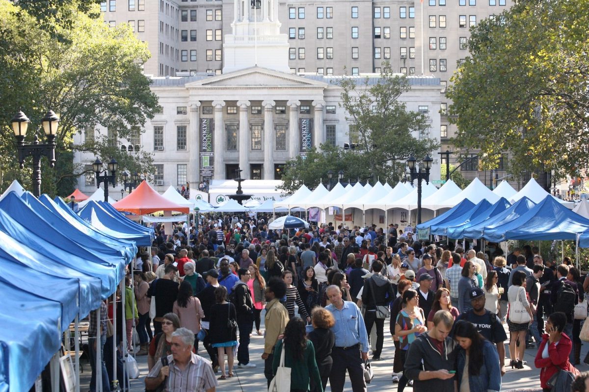 A large crowd of people stand and walk around by blue, white and red tents. Behind the tent area is a white building with two banners that read “Brooklyn Book Festival.”