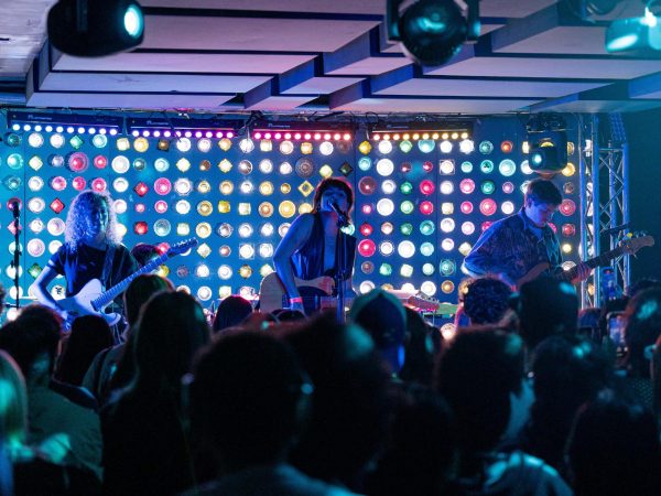 Singer-songwriter and N.Y.U. student Val Olson, or Deadbeat Girl, sings in front of a crowd while holding a guitar. On her left stands a guitarist, and on her right stands a bass player.