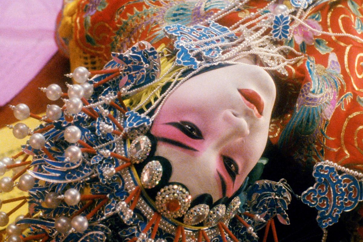 An upside-down shot of actor Leslie Cheung, his face covered in white face paint, and pink-and-black makeup. He is wearing an orange-and-blue, jewel-covered outfit, a matching headpiece and pearls.