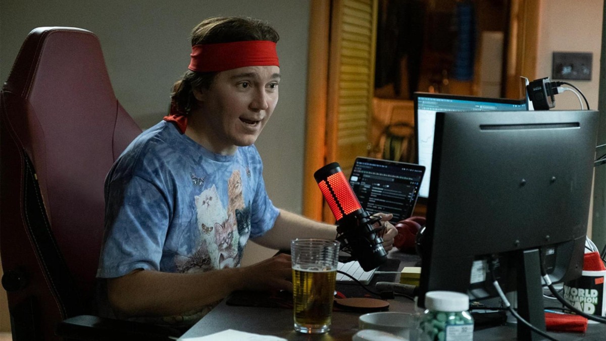 Actor Paul Dano wearing a blue shirt and red headband sits in a brown desk chair. He is speaking into a red microphone while looking at two computer monitors. The image is from the film “Dumb Money.”