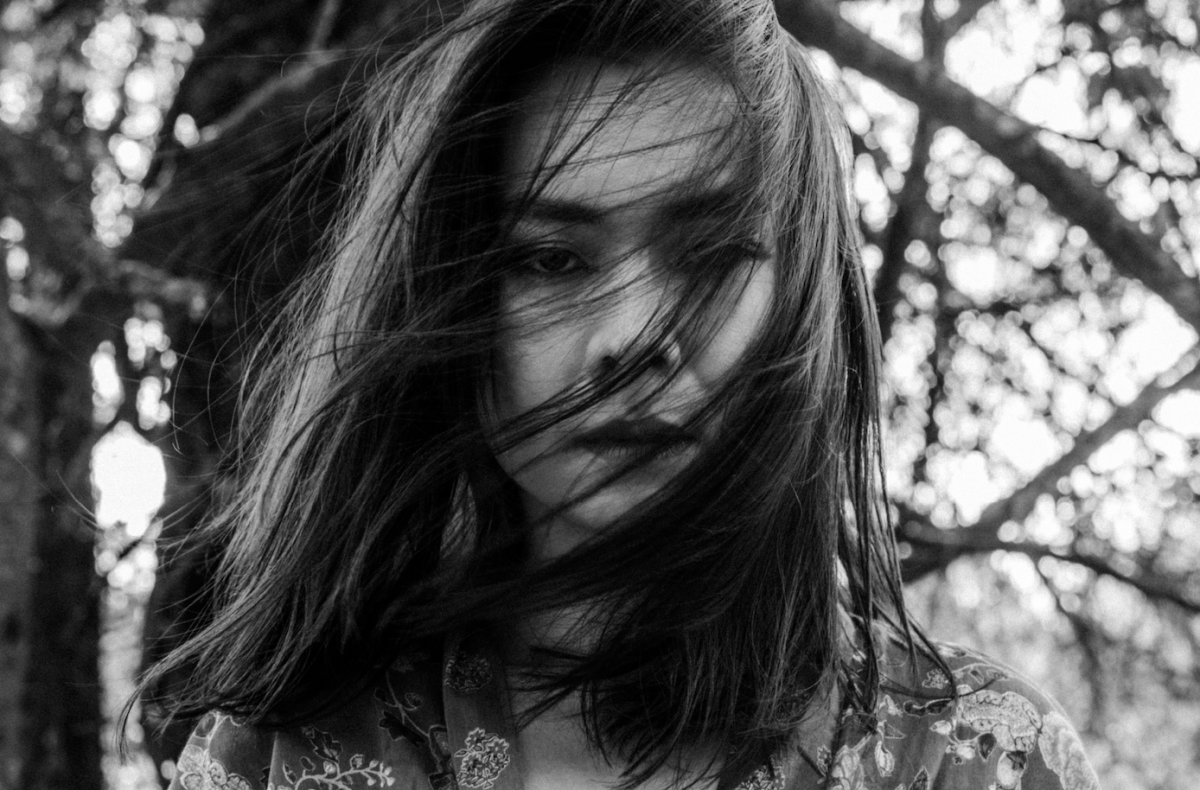 Singer-songwriter Mitski with dark, shoulder-length hair windswept across her face stares into the camera in front of many tree branches. The photo is black and white.