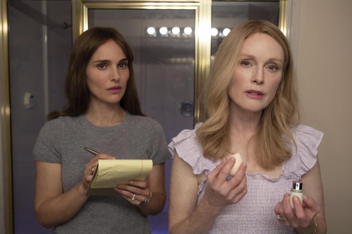 Actresses Natalie Portman, on the left, and Julianne Moore, on the right, look into the camera as if it is a mirror. Natalie Portman holds a notepad and pen while Julianne Moore holds a makeup sponge and lipstick. The image is from the film “May December.”