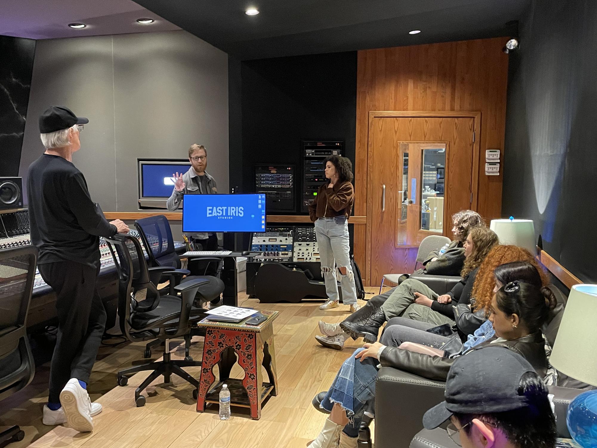 A group of students sitting in a music recording studio while listening to two instructors speaking. The room is filled with various music production equipment.