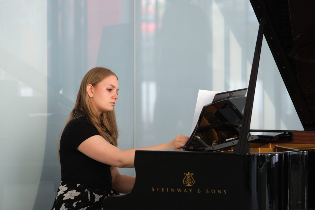 A girl with dark blonde hair and a black top is sitting in front of a piano, adjusting the sheet in front of her. It says, with gold letters, ‘Steinway and Sons,’ on the piano.