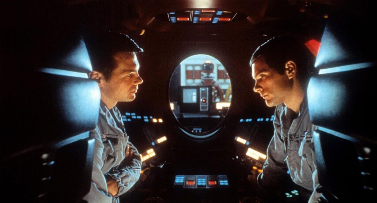 Two men lean forward and stare at each other in a spaceship. There is a round window in the center and orange, blue and red buttons behind them.
