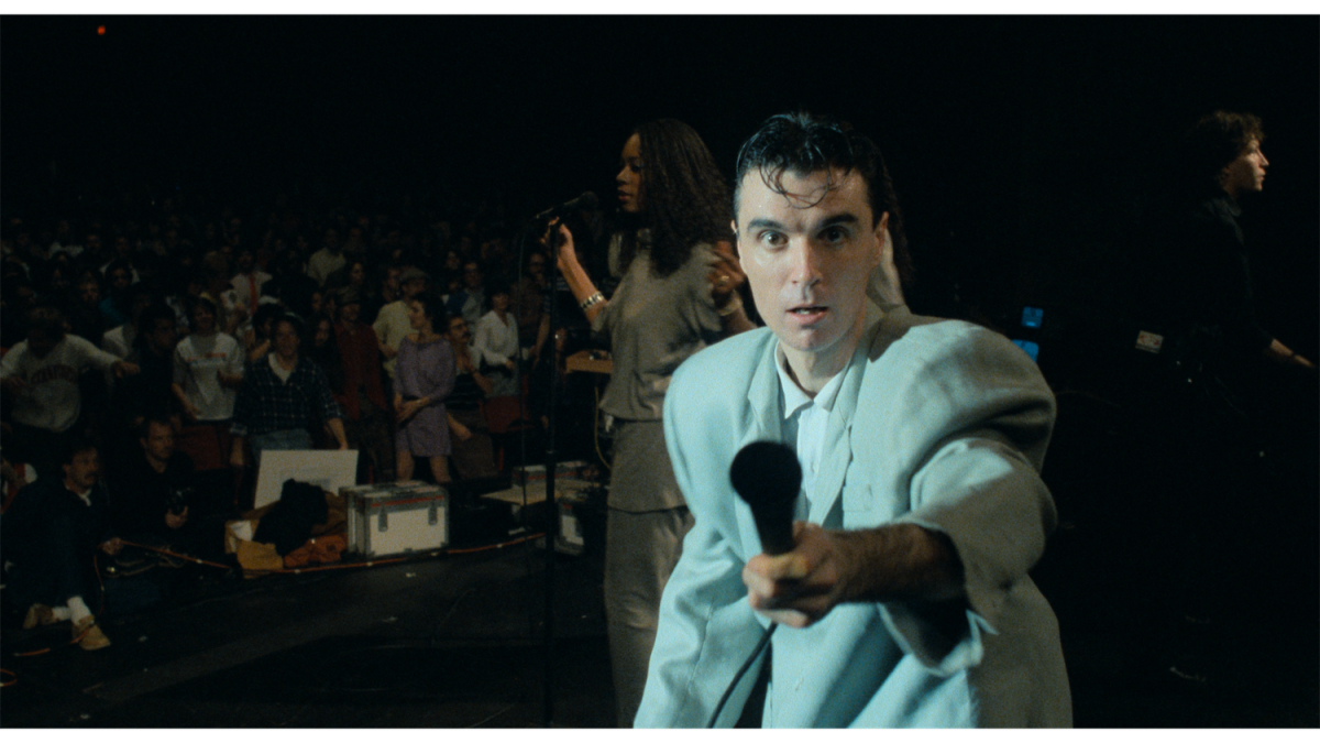 A still image from the film “Stop Making Sense” with lead vocalist David Byrne singing on stage and pointing a microphone to the camera.