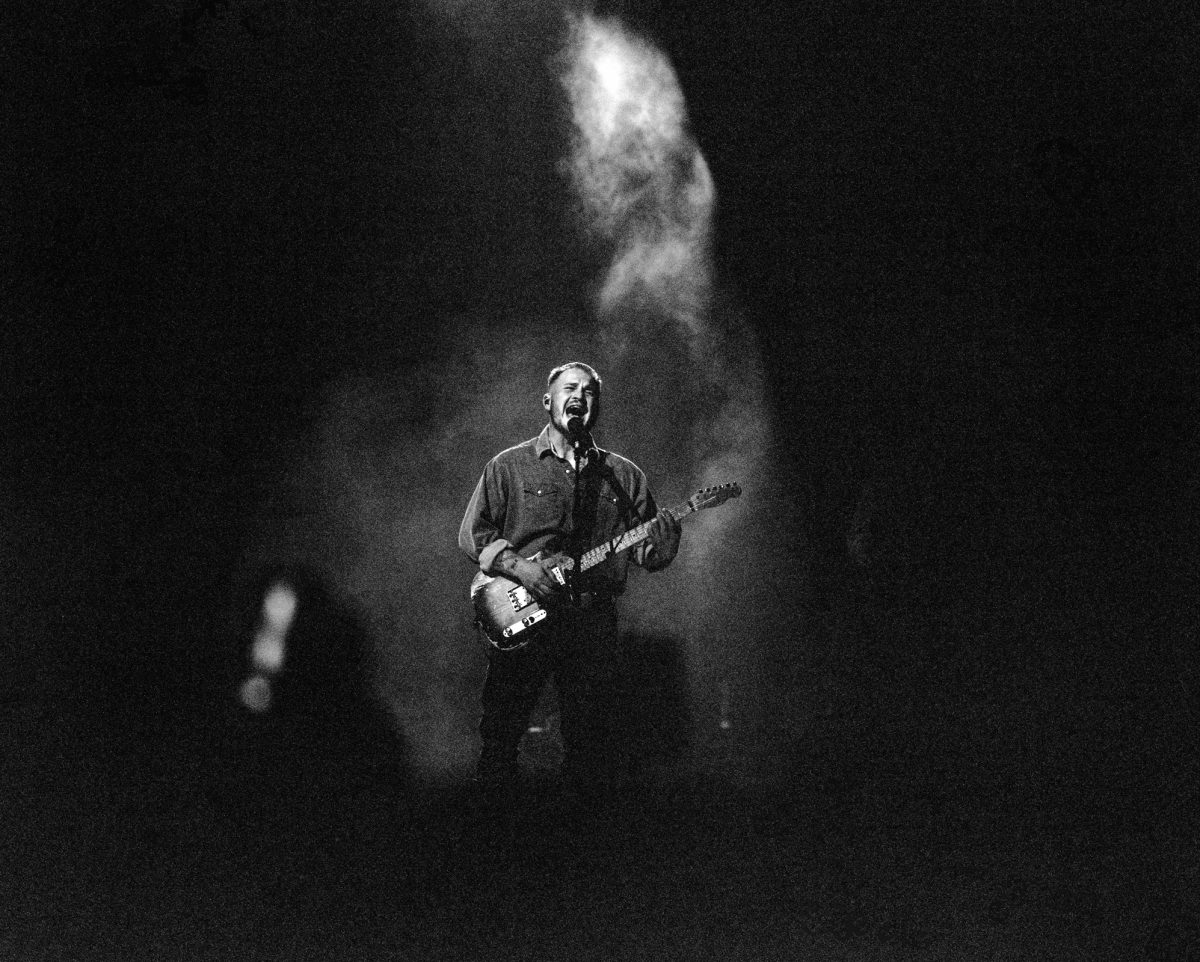A black and white shot of singer-songwriter Zach Bryan. He is standing behind a microphone and singing while playing the guitar. There is smoke going up behind him.