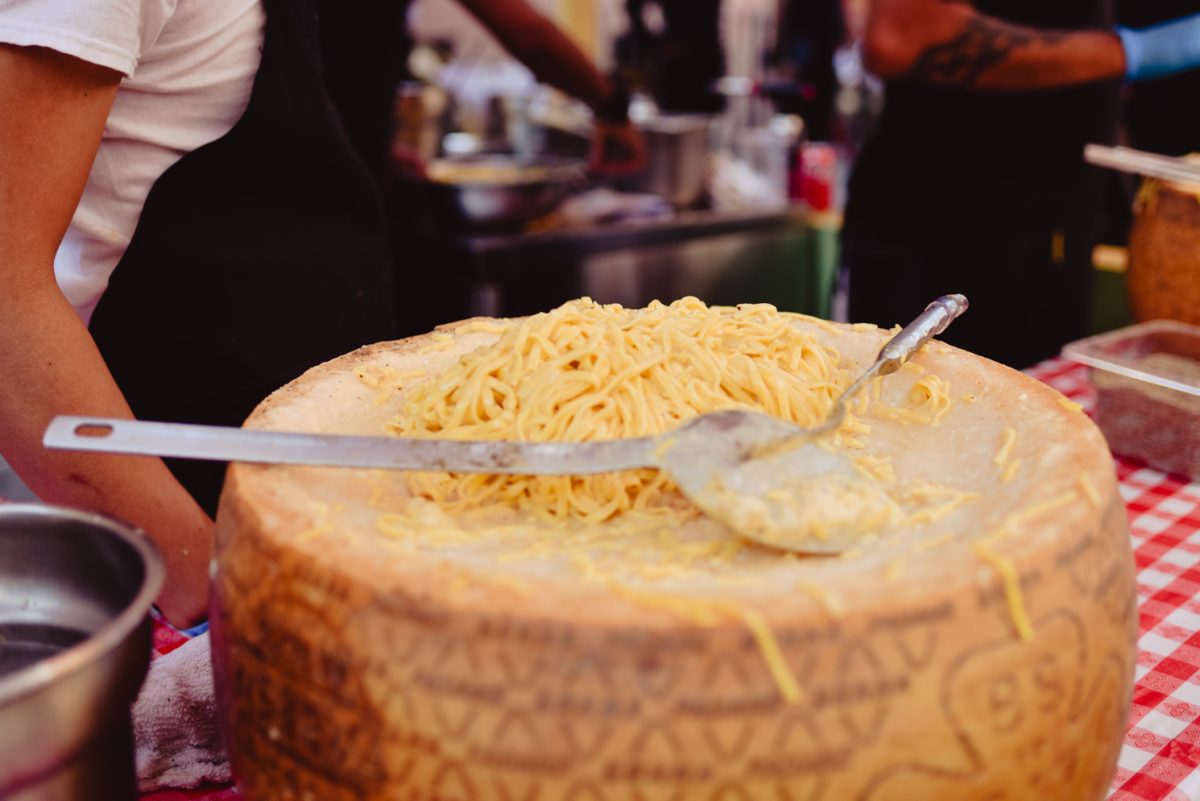A close-up shot of pasta Alla Ruota on a pale yellow cheese wheel. A large, silver spoon sits on top of the wheel. The chest and right arm of a person wearing a black apron is visible behind the wheel, which sits on a table covered with a red-and-white checkered tablecloth.
