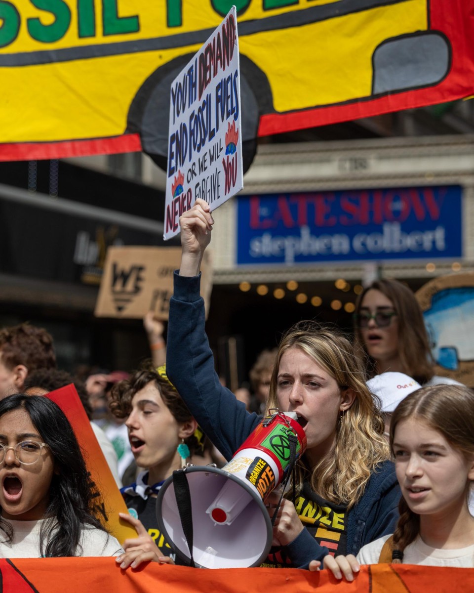 In a crowd, a blonde woman raises her hand holding a poster in one hand and a red-and-white megaphone in the other.