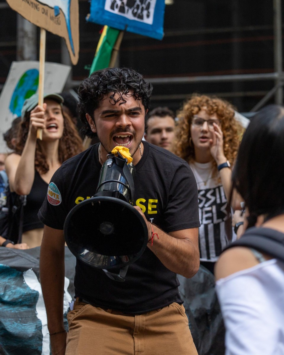 A man wearing a black shirt and brown pants holds a black megaphone with his left hand. He is speaking into it.