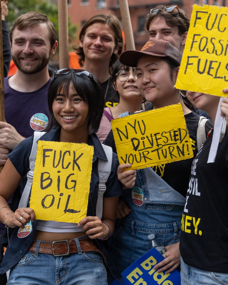 A group of seven people pose with yellow signs that read in black colored marker: “Fuck Big Oil”, “N.Y.U Divested Your Turn” and “Fuck Fossil Fuels”.