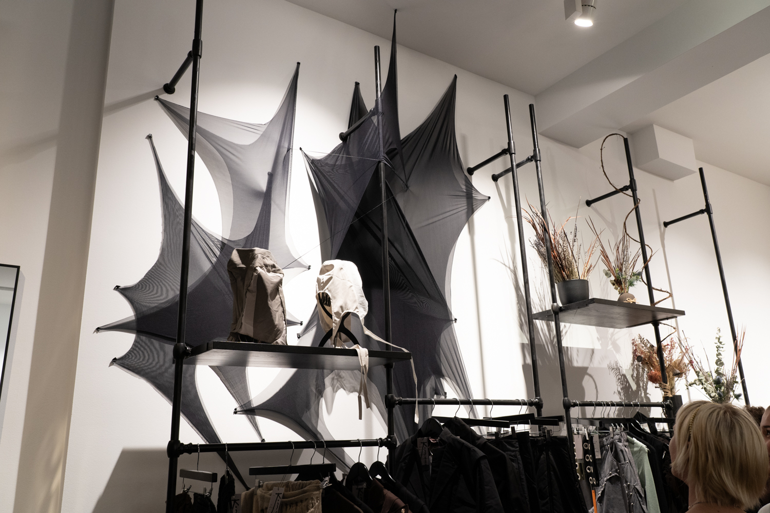 A collection of jackets, trousers and hats hanging under a shelf. The top of the shelf is decorated with dark clothes and plants.