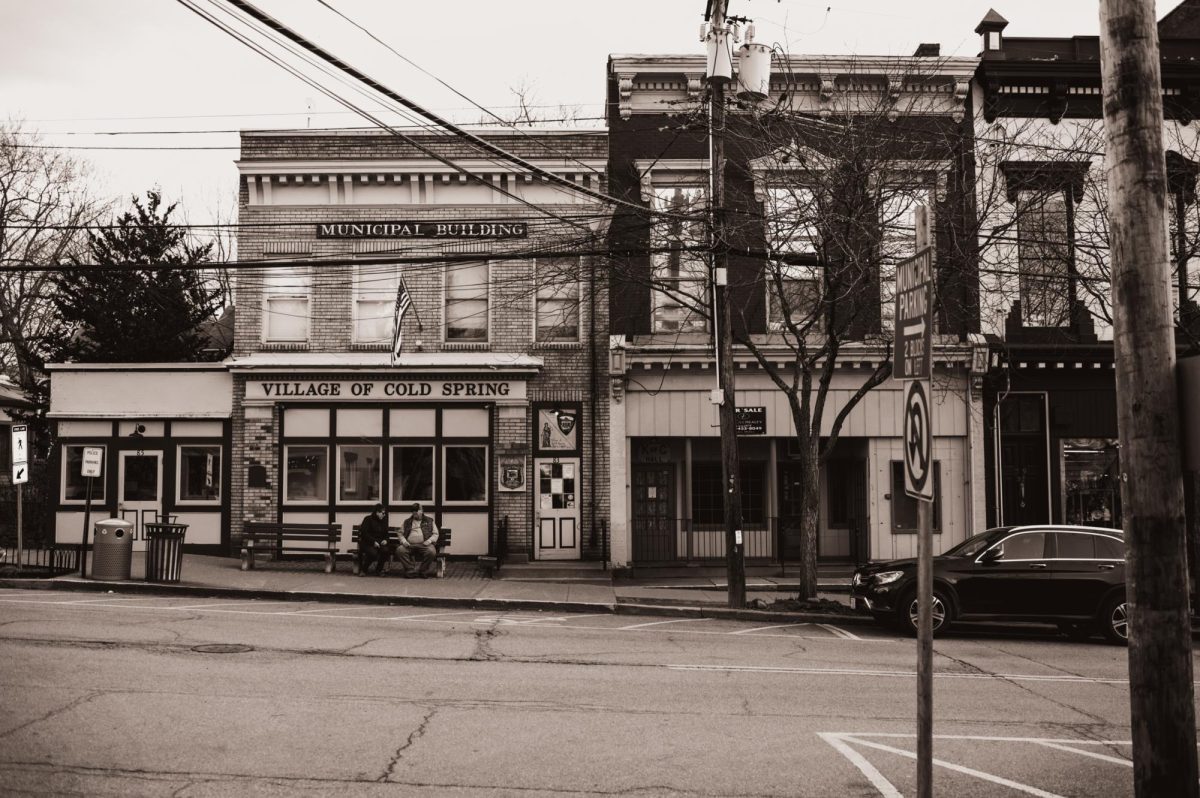 A black and white photo showing the side of a road in Cold Spring, New York. Two people are sitting on a bench in front of a building.