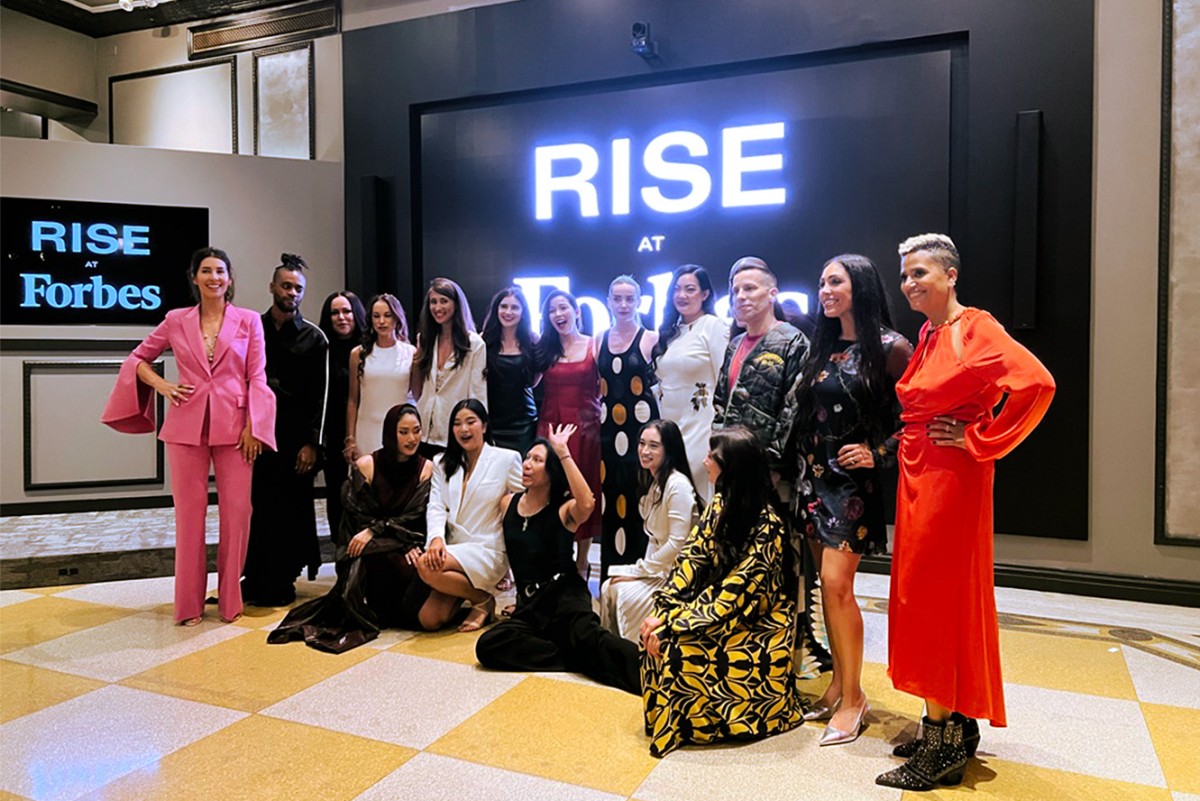 A group of attendees wearing various dresses pose for a photo in front of a sign that reads ” Rise at Forbes.”
