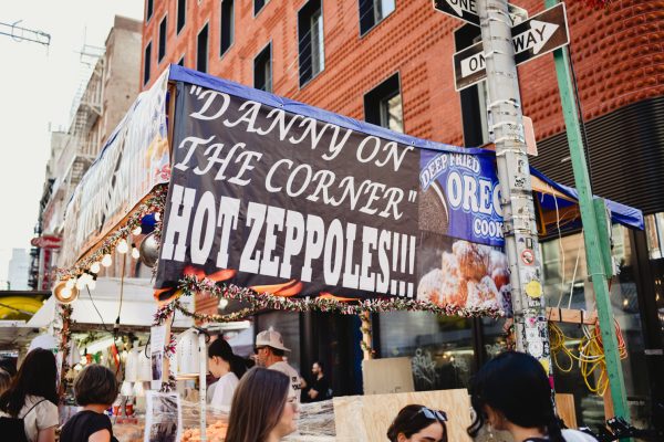 The side of a store that has a sign which reads “DANNY ON THE CORNER HOT ZEPOLLES” followed by three exclamation points. To the right of it is a blue sign showing deep-fried Oreo cookies. There are people standing around the store.