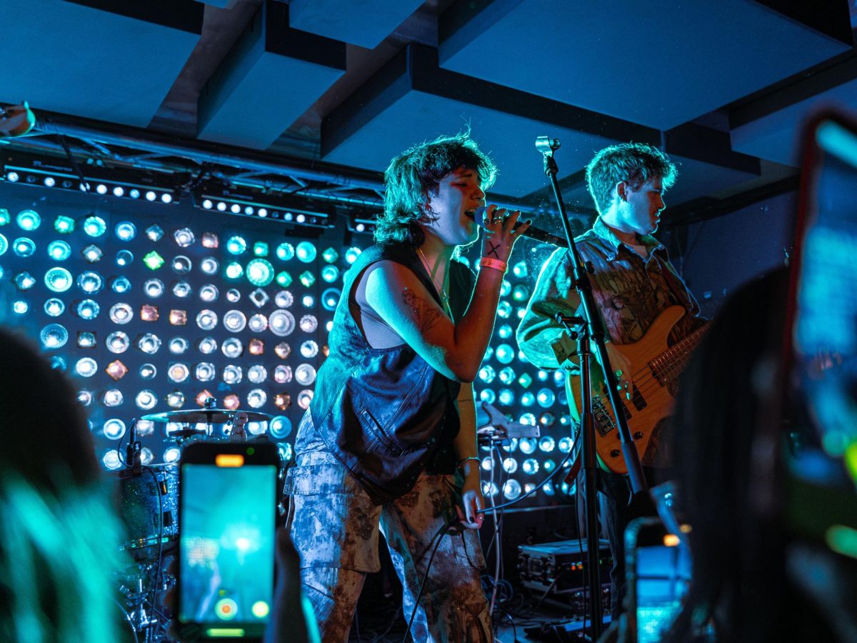 Singer-songwriter and N.Y.U. student Val Olson, or Deadbeat Girl, sings on a concert stage lit up in blue alongside a bass player. Under the stage, audience members record her performance on their phones.