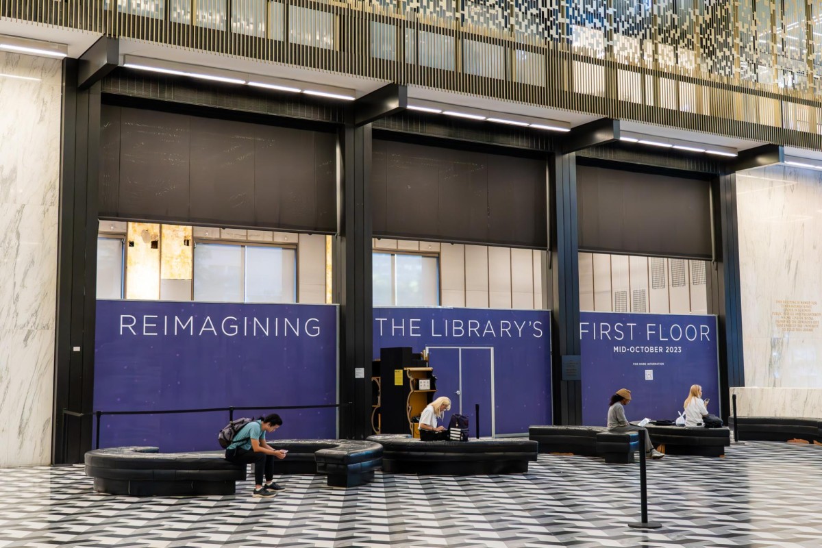 Four people sit in the first floor of Bobst Library with purple construction wallpaper with the text REIMAGINING THE LIBRARY’S FIRST FLOOR” behind them.