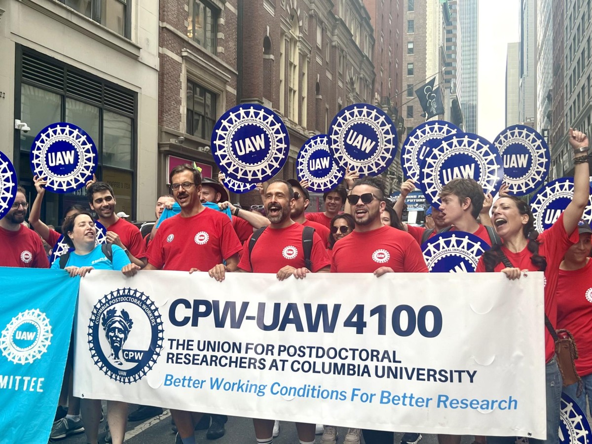A group of UAW members wearing red and blue shirts hold up a sign reading “CPW-UAW 4100 The Union for Postdoctoral Researchers at Columbia University”.