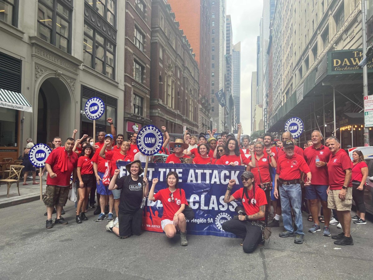 A photo of N.Y.U. Contract Faculty United and Researchers United protesting, wearing red shirts and holding blue U.A.W. signs and a blue banner.