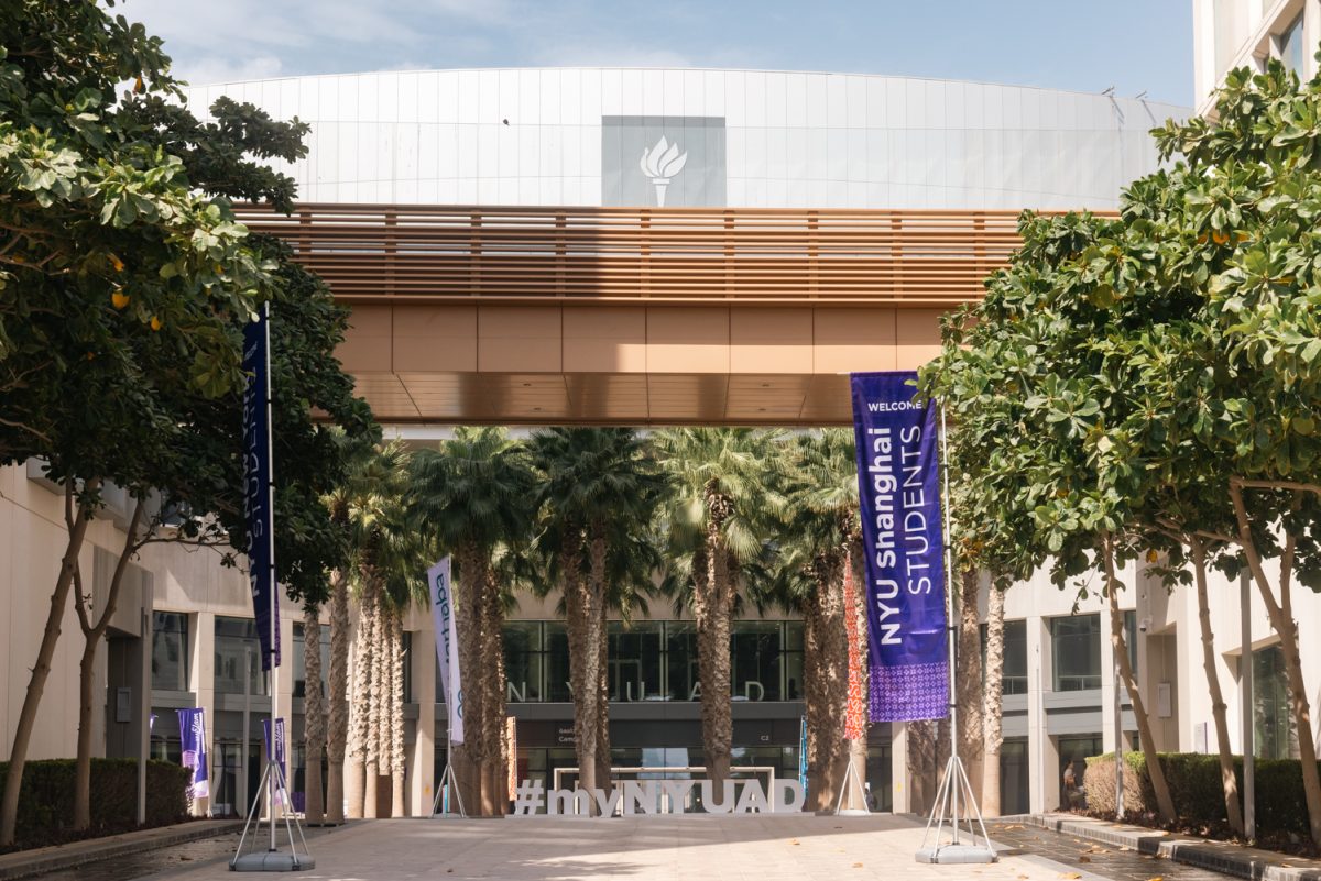The front view of an N.Y.U. Abu Dhabi building with a bridge across the row. Numerous rows of palm trees stand in front of the building alongside two purple flags.