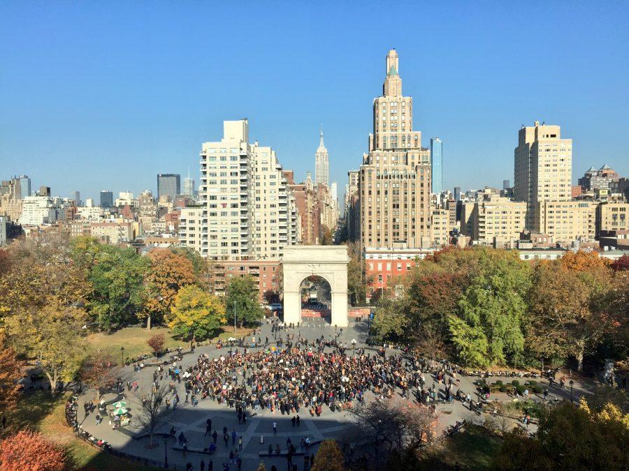 An aerial view of the Washington Square Park and the Midtown skyline. Crowds gather around the fountain in the park. The white Washington Square Arch is in the center of the image.