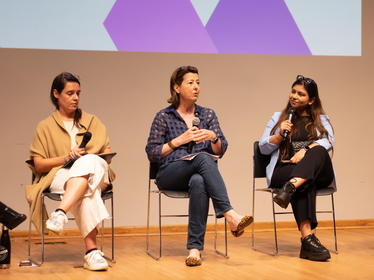 The dean of N.Y.U.’s Tandon School of Engineering, Jelena Kovačević, speaks into a microphone on a stage. Two people are sitting next to her on either side.