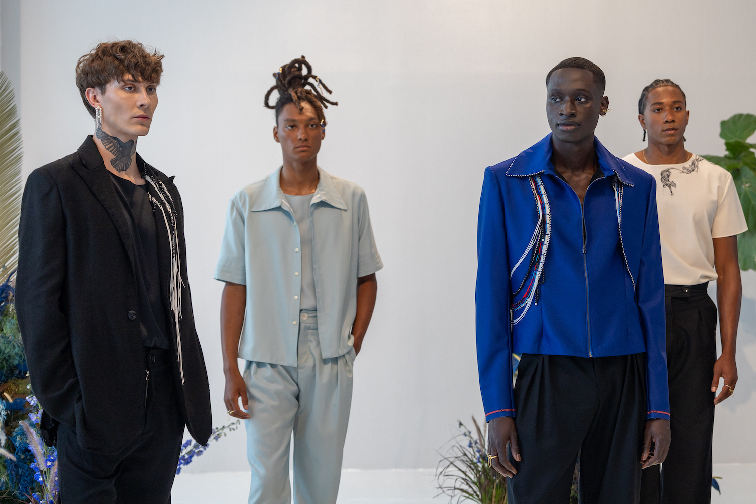 Four models stand in a white showroom with floral plants in the background. The models are wearing clothes from KENT ANTHONY.