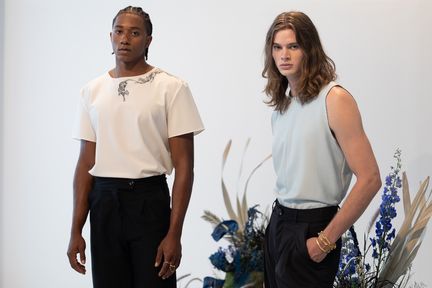 Two models in the center of the room stand with floral decorations in the background. The models are wearing clothes from KENT ANTHONY.