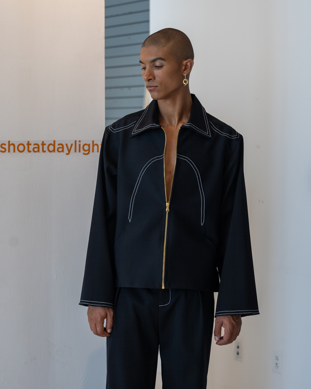 A model wearing a black jacket with gold hardware looks down. The model is wearing clothes from KENT ANTHONY.