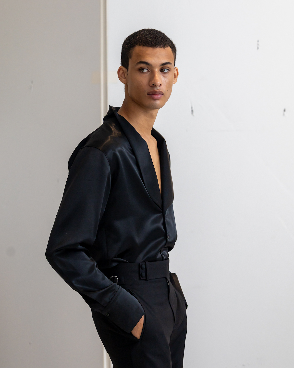 A model wearing a black suit leans against a wall. The model is wearing clothes from B.M.C.