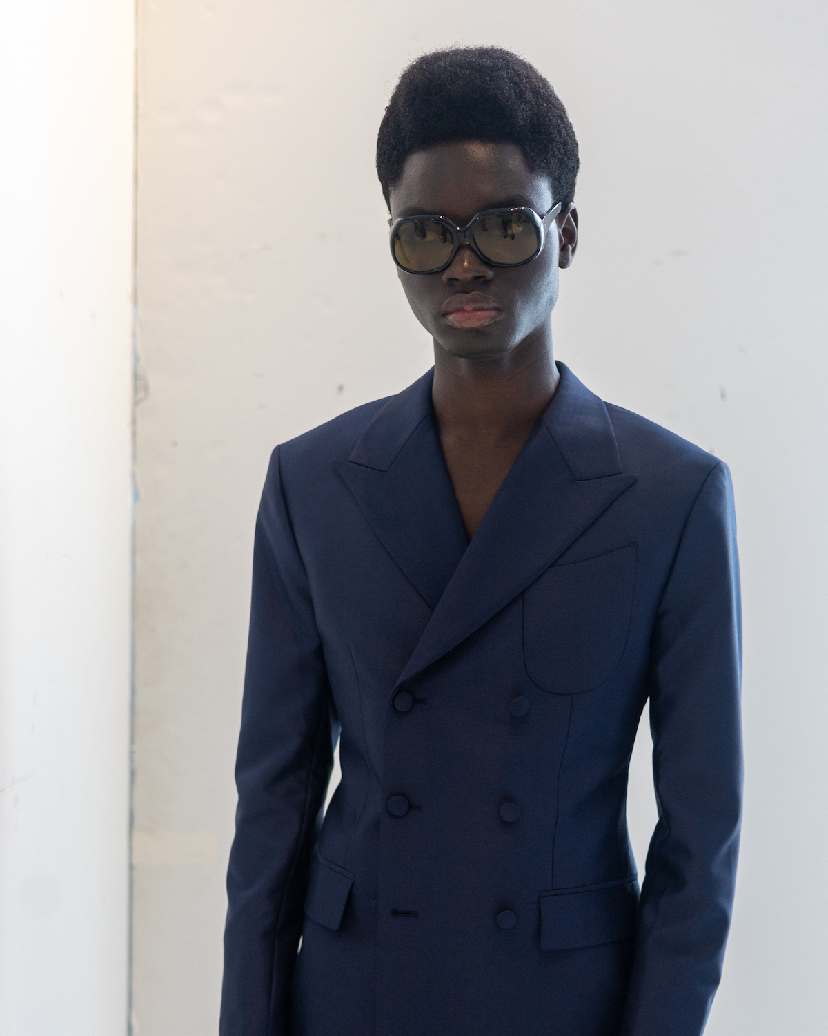 A model wearing black sunglasses and a navy suit stands in a white showroom. The model is wearing clothes from B.M.C.