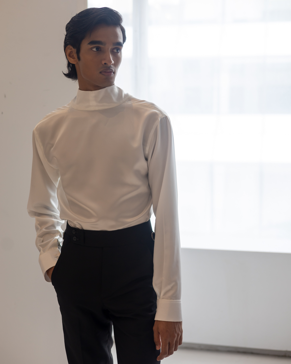 A model wearing a white long-sleeved turtleneck and black suit pants stands in a white showroom. The model is wearing clothes from B.M.C.