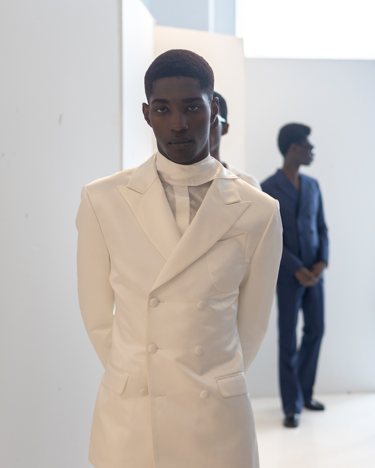 A model wearing a white suit stands in a white showroom. The model is wearing clothes from B.M.C.