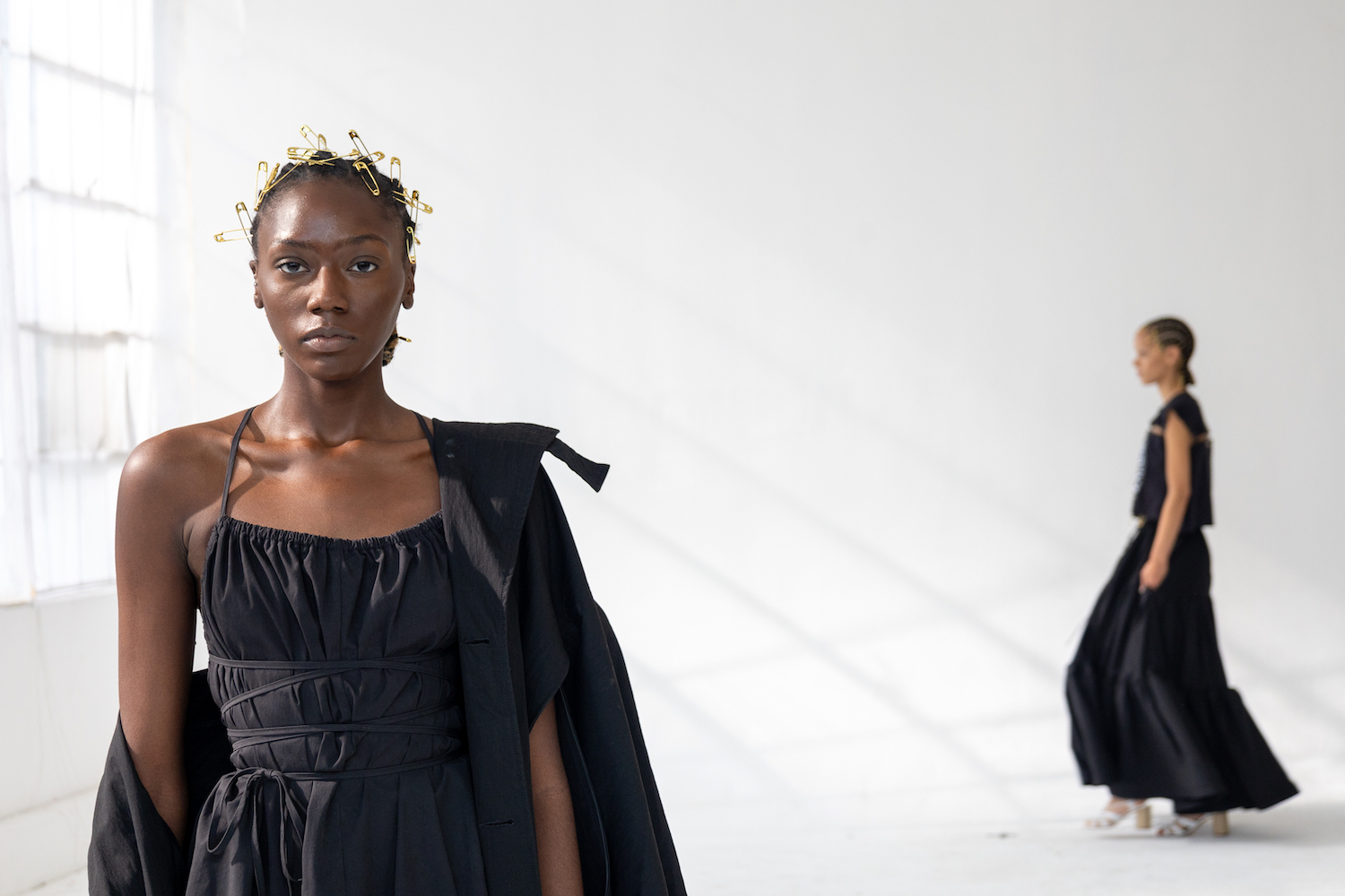 A model wearing a black dress with a gold headpiece walks down a short runway. The model is wearing clothes from A.POTTS.