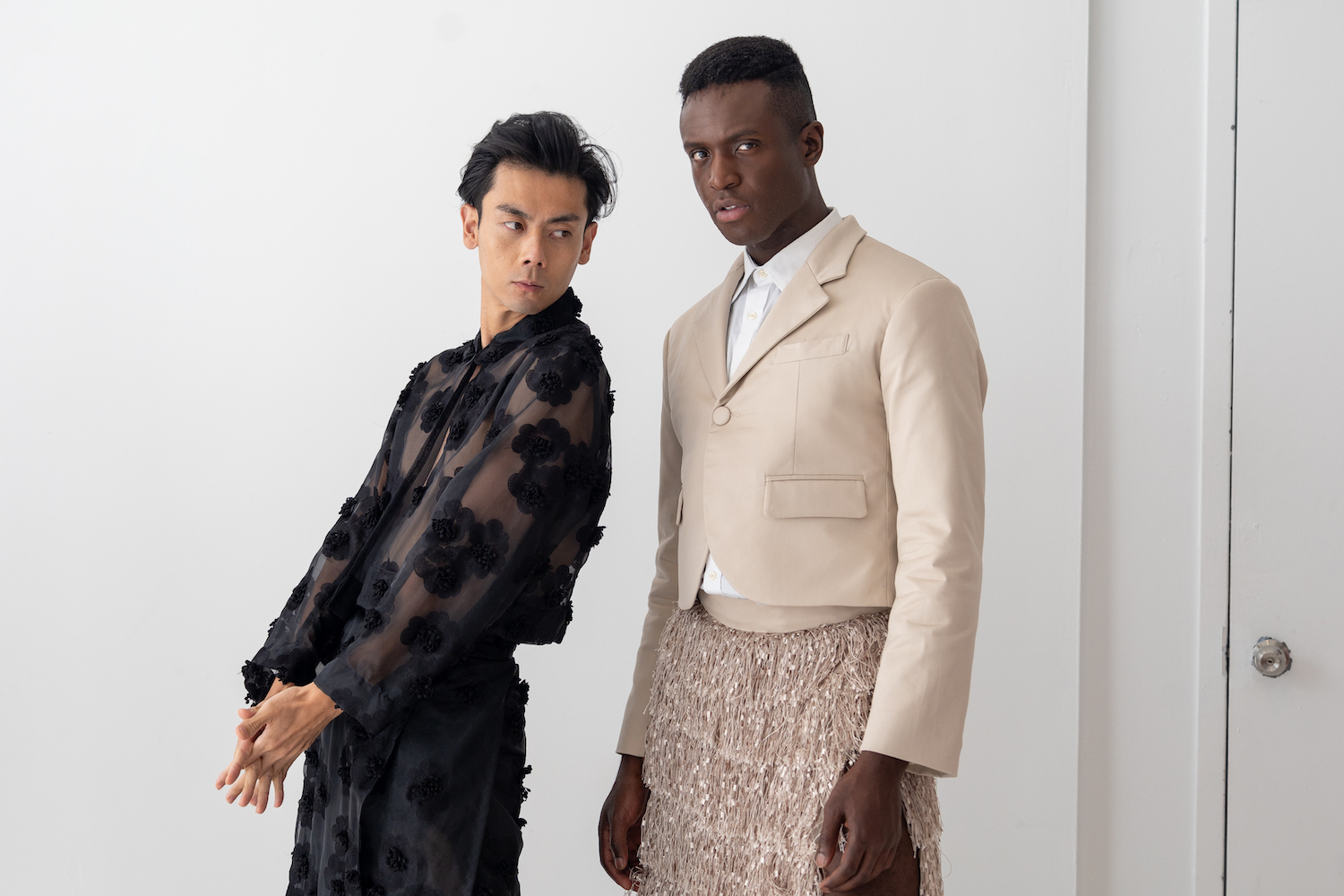 Two models pose together near a wall of the showroom. One model is wearing a black mesh top, and the other model is wearing a cream blazer. The models are wearing clothes from TERRY SINGH.