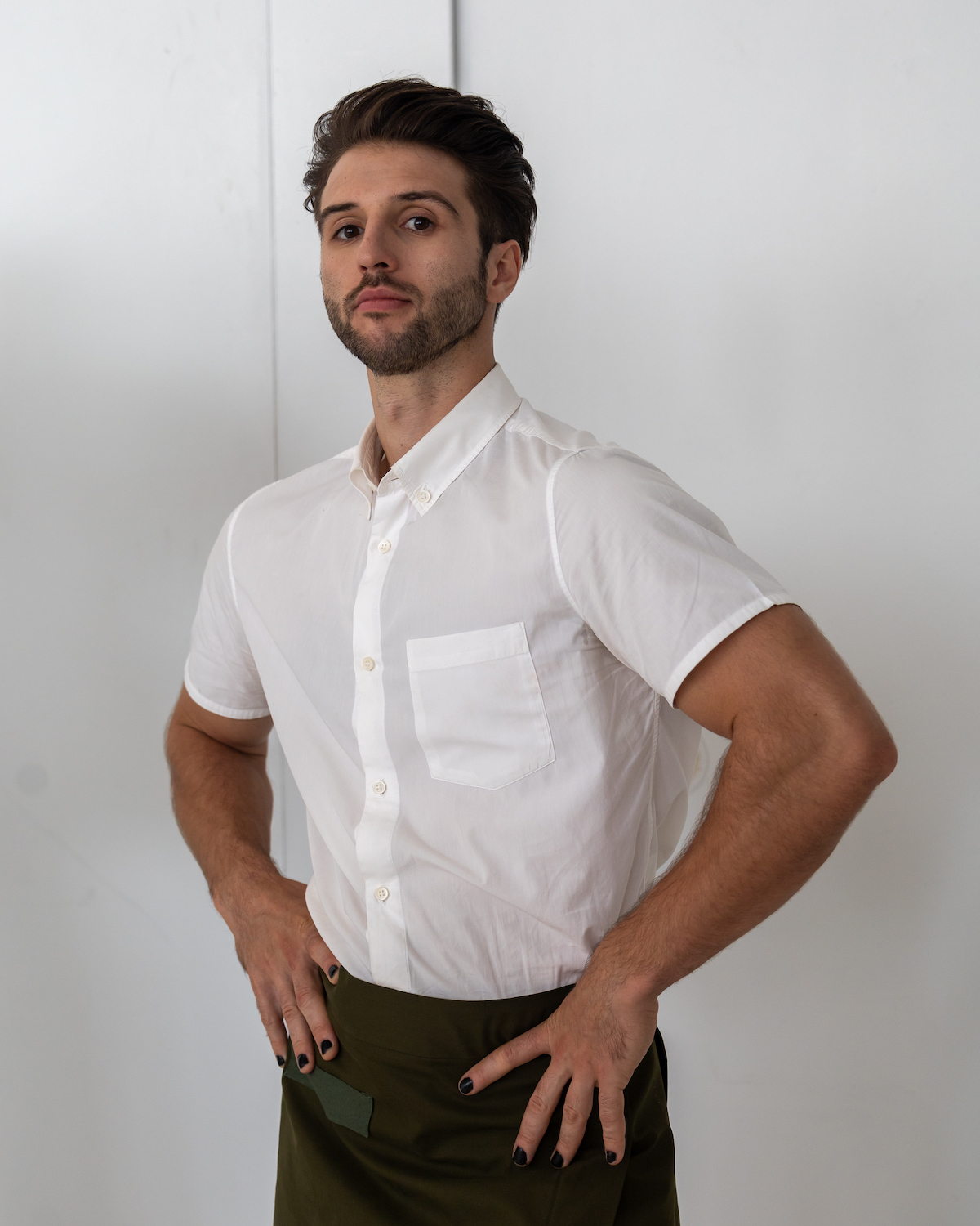 A model wearing a white shirt and dark green skirt poses with their hands on their hips. The model is wearing clothes from TERRY SINGH.