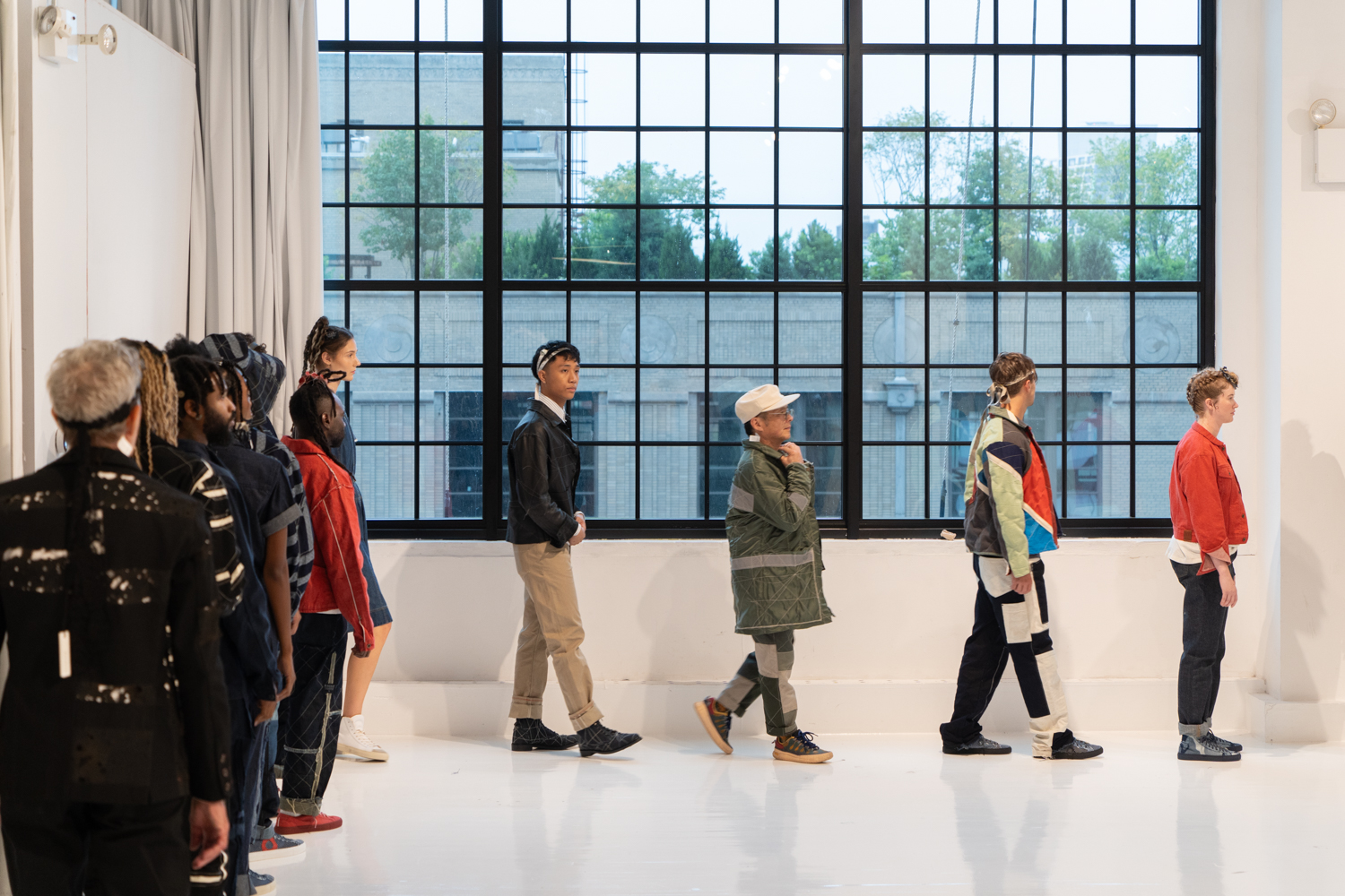 A line of models walks across a fashion showroom with white walls and floors. They are wearing clothing from the brand Raleigh Workshop.