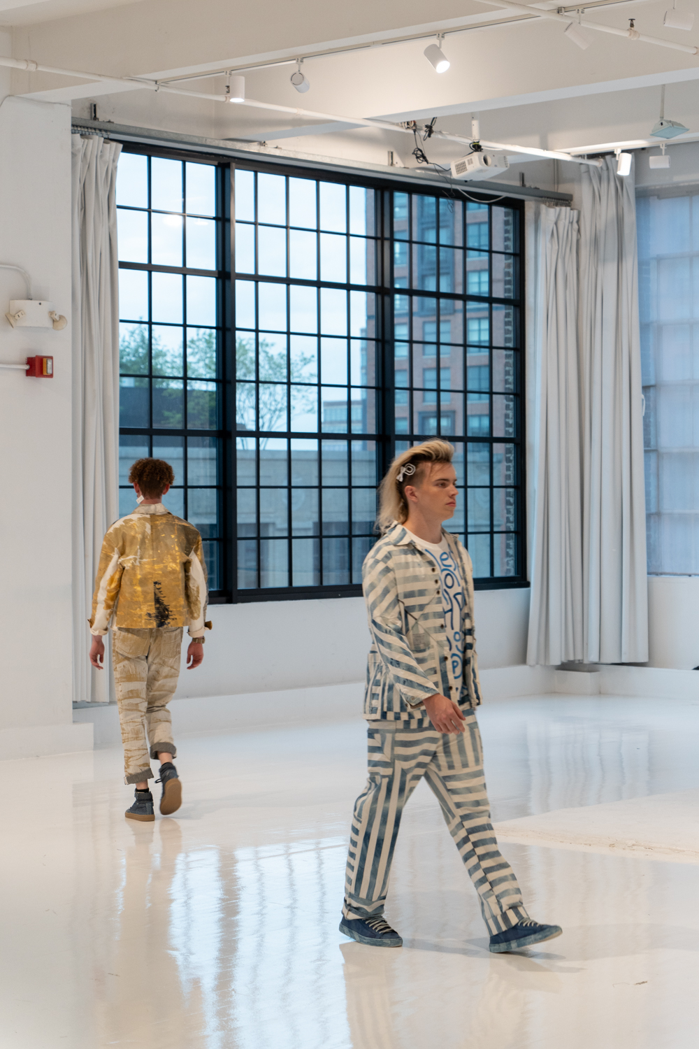 Two models walk across a fashion showroom with white walls and floors. They are wearing clothing from the brand Raleigh Workshop.