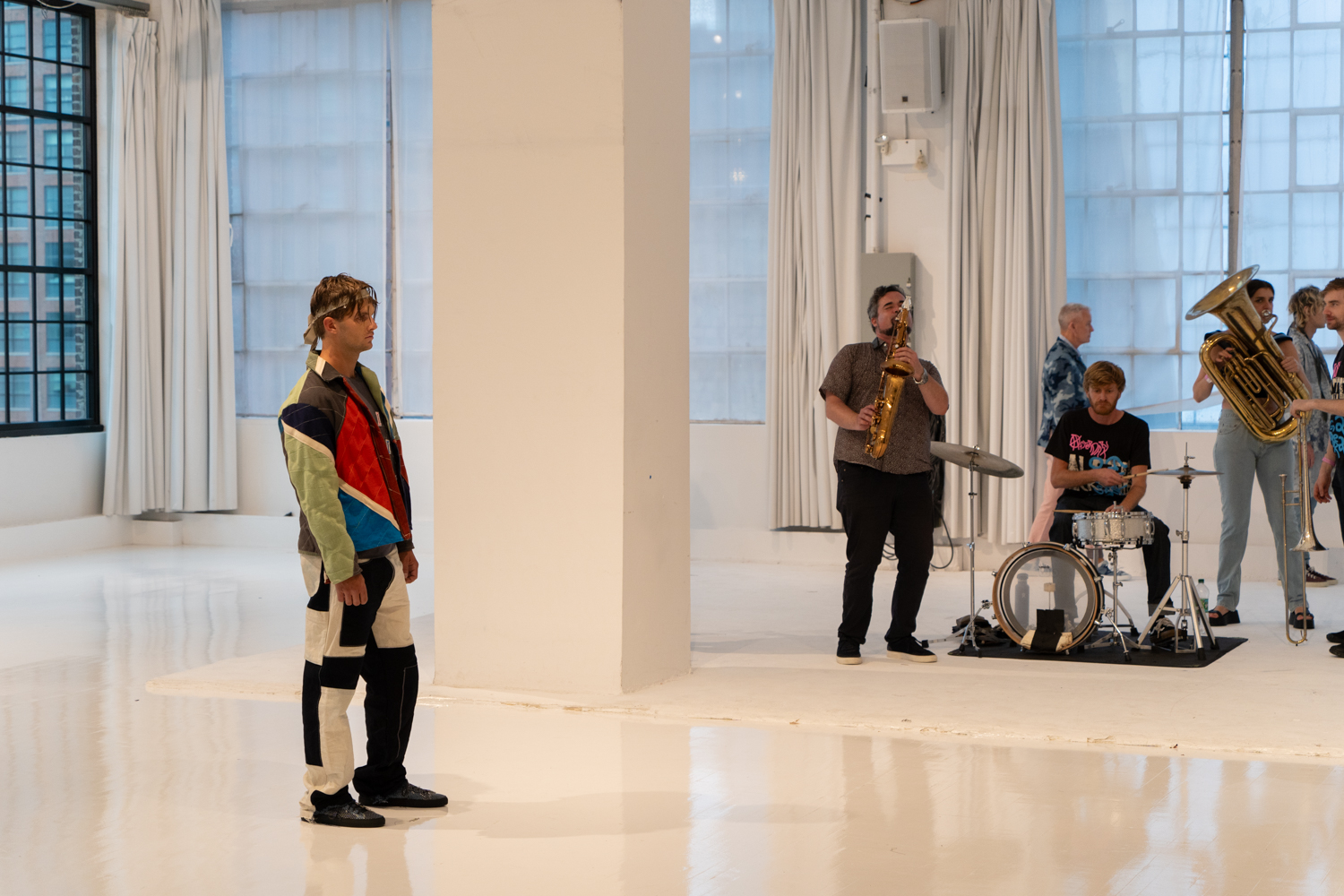 A model poses in the middle of a fashion showroom with white walls and floors and a band is playing. They are wearing clothing from the brand Raleigh Workshop.