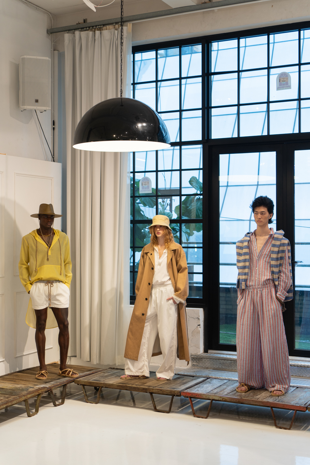 Three models stand in a fashion showroom with white walls and floor-to-ceiling windows. They are wearing clothing from the brand thesalting and are standing on wooden cargo racks.