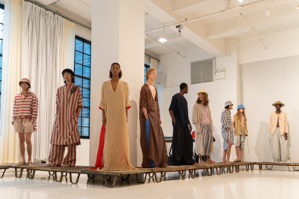 Nine models stand in a fashion showroom with white walls. They are wearing clothing from the brand thesalting. They are standing on wooden cargo racks placed on the floor across the room.