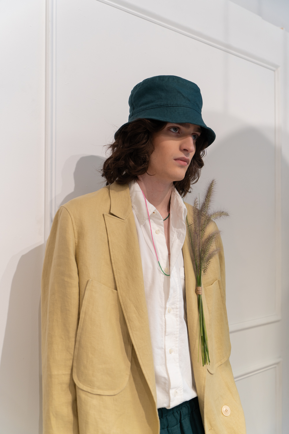 A model stands in a fashion showroom with white walls. The model is wearing a green bucket hat, a beige suit jacket, a white shirt and green pants from the brand thesalting. There is a small bouquet of foxtail attached to the jacket.