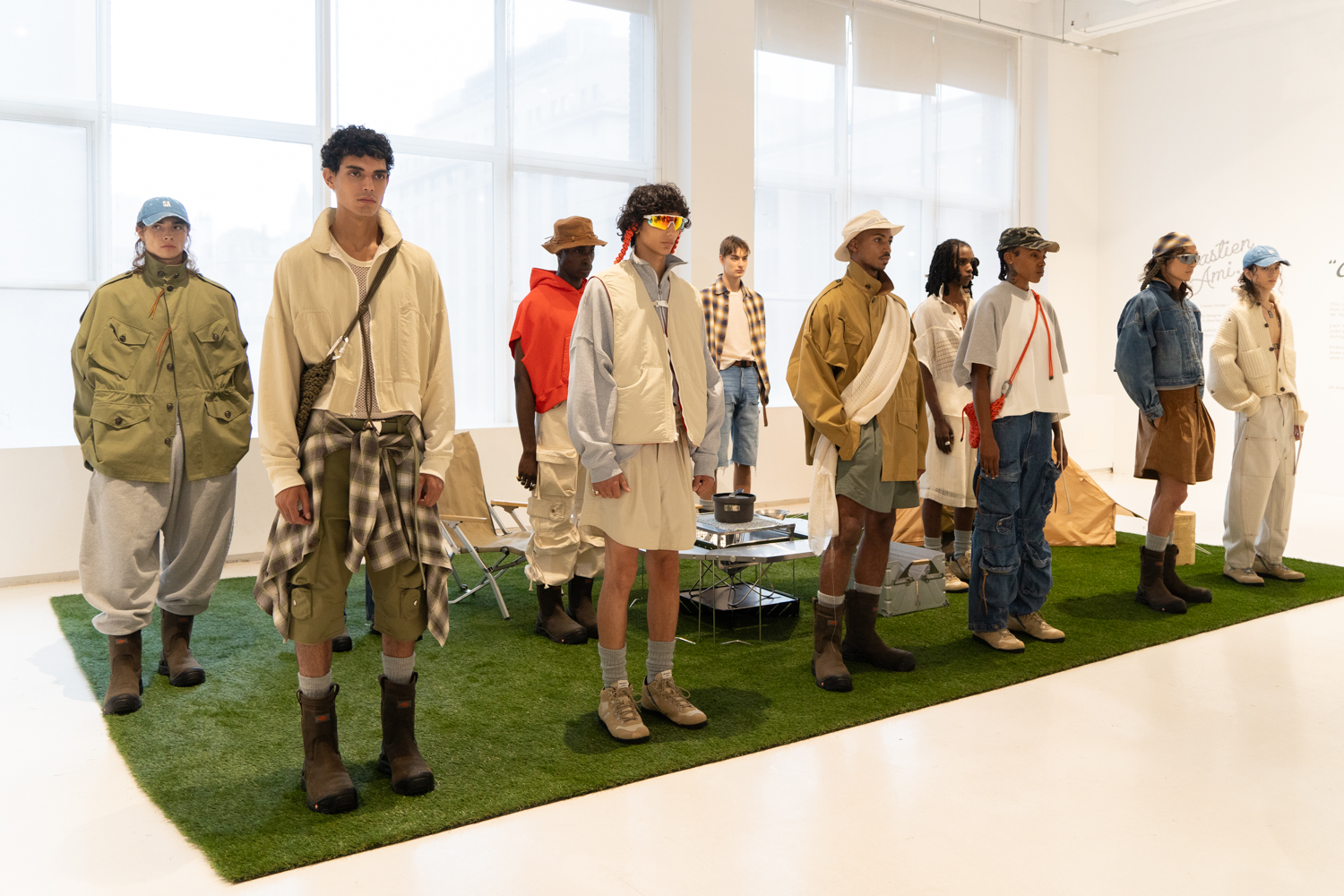 Models stand in two lines in a fashion showroom with white walls and floors. They are standing on a carpet of artificial turf with camping equipment. They are wearing clothing from the brand SEBASTIEN AMI.
