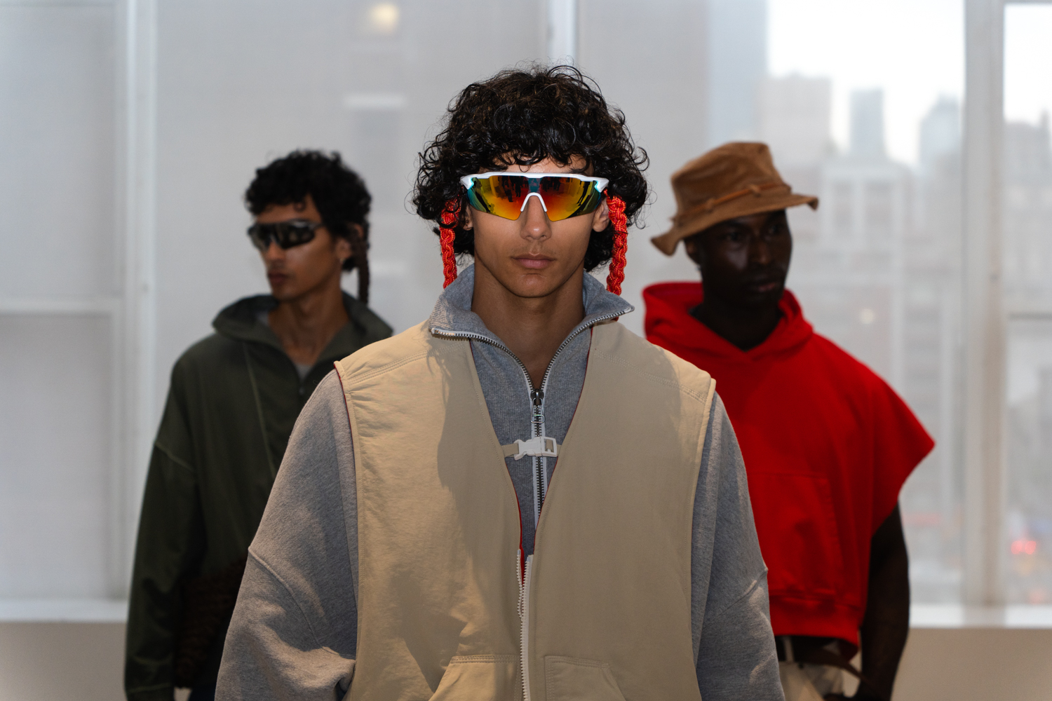 A model wearing reflective sunglasses, a gray jacket and a beige vest stands in a fashion showroom. Behind the model are two other models. They are wearing clothing from the brand SEBASTIEN AMI.