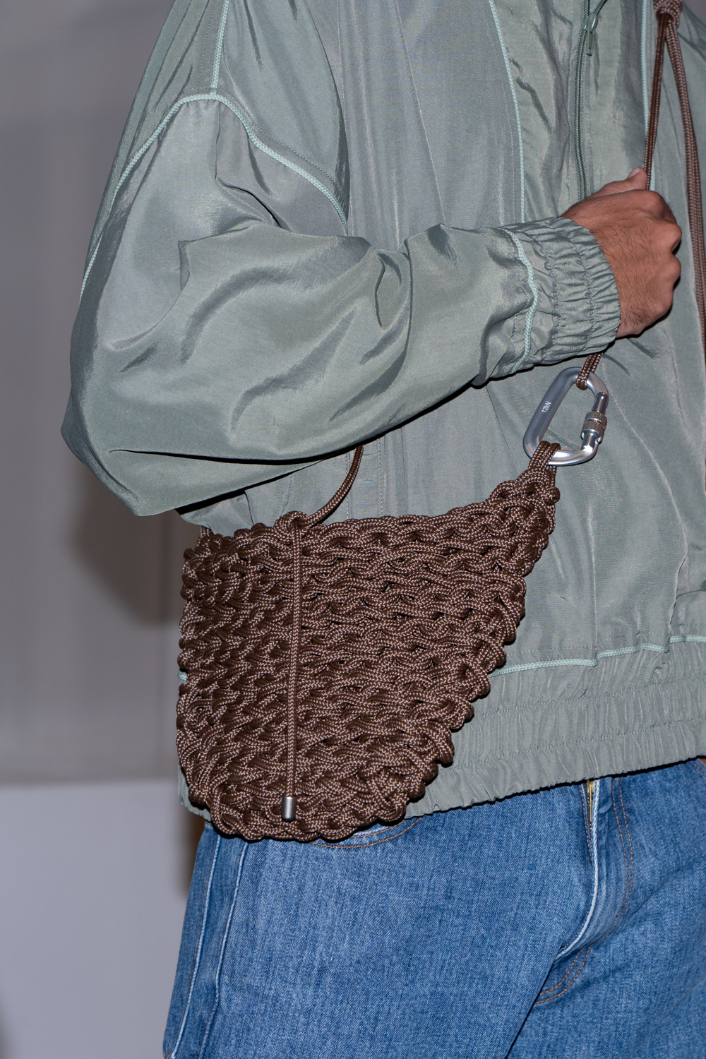 A close-up photo of a model wearing a green jacket and blue jeans. The model is holding a brown knitted bag from the brand SEBASTIEN AMI.