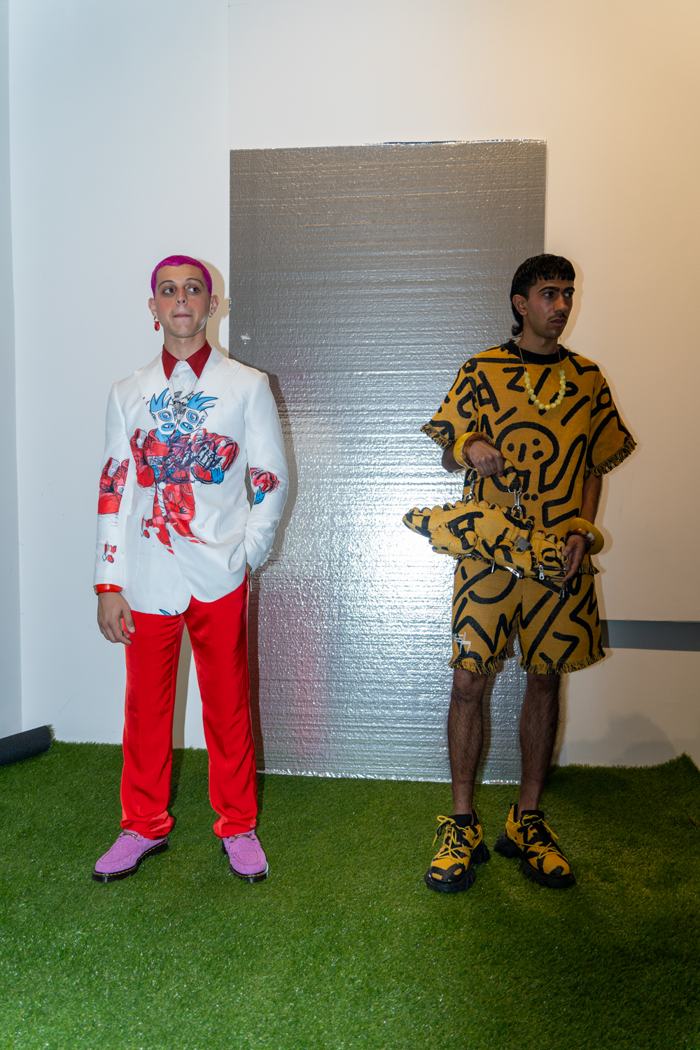 Two models stand in a fashion showroom with a reflective piece of cardboard behind them and artificial turf on the floor. They are wearing clothing from the brand Skyco.