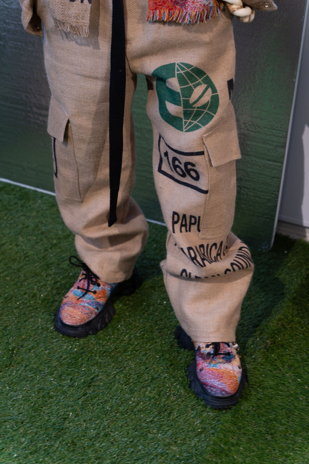 A close-up photo of a model wearing beige nylon pants and boots with colorful patterns. The model is standing on artificial turf and wearing clothing from the brand Skyco.